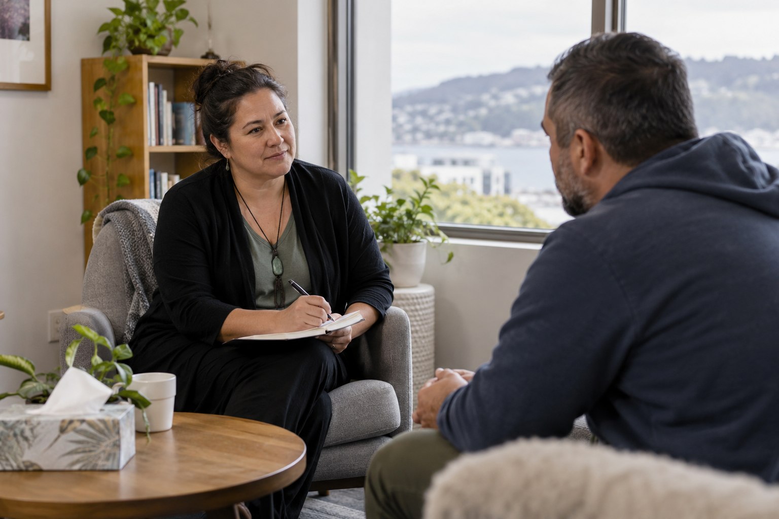 A support counsellor speaking with an adult client in a calm neutral consultation room, respectful medium-wide documentary view