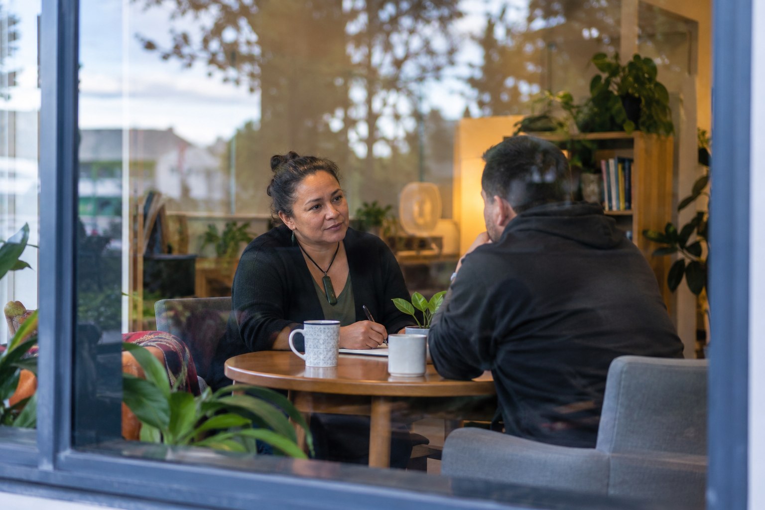 A support worker listening to an adult client across a small table in a community wellbeing room, unposed and respectful